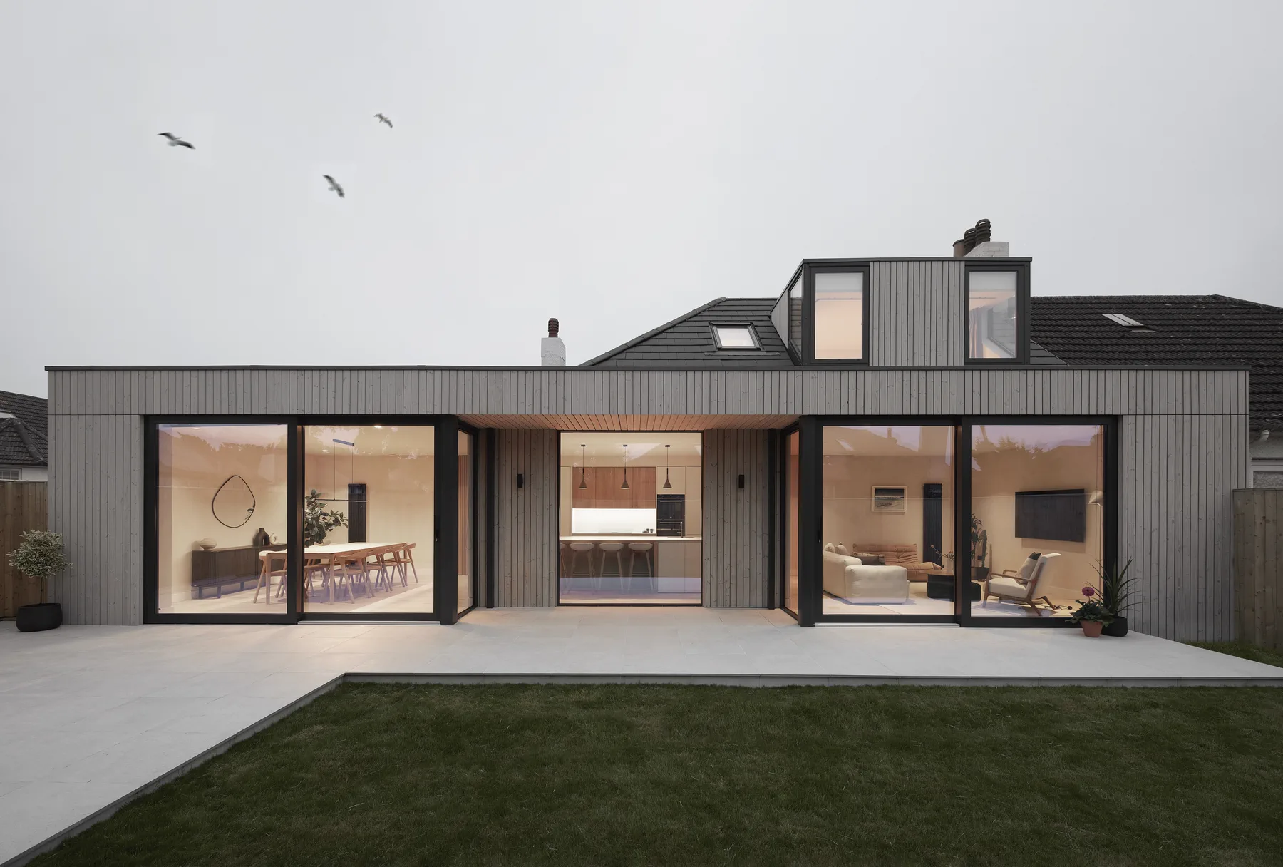 A single-storey extension clad in vertical Thermal Pine rainscreen, weathered silver-grey, with full-height glazing onto a lawn.